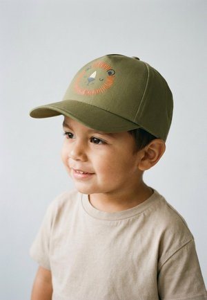 Kleines Kind trägt eine olivgrüne Mütze mit einer Illustration eines lachenden Löwengesichts und ein beiges T-Shirt, blickt leicht zur Seite und lächelt.