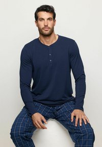 Námořnická modrá dlouhá košile henley s tříknoflíkovým zapínáním, spárovaná s modrými kostkovanými pyžamovými kalhotami s bílými pruhy. Hladká textura, uvolněný střih.