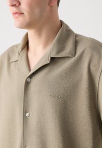 Beige textúrájú ing, kivágott gallérral, elöl gombokkal és finom „HUGO” logóval, azonos színben. Puha anyag, laza szabással.