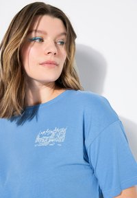 Blauwe katoenen t-shirt met een witte grafische afbeelding die lijkt op een waterfrontscène op het linkerborstgebied. Korte mouwen, relaxed fit.