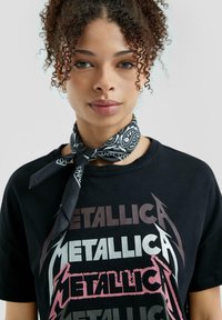Zwarte t-shirt met een gelaagd "Metallica"-grafiek in tinten grijs, roze en zilver. Gecombineerd met een zwart-wit gestreepte sjaal om de nek.