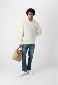 Suéter beige, camisa de cuadros azul, jeans azules y zapatillas blancas. Sosteniendo un bolso color canela con cremallera y asas marrones. Atuendo casual.