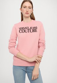 Rozā džemperis ar uzrakstu "VERSACE JEANS COUTURE" treknā melnā burtu stilā. Izstrādāts ar apaļu kakla izgriezumu un ribota raksta aprocēm, izgatavots no mīksta auduma.