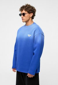 Blauwe sweatshirt met een ronde hals, oversized pasvorm en ribgebreide boorden. Kenmerkt zich door een klein wit logo op de borst. Zwarte broek.