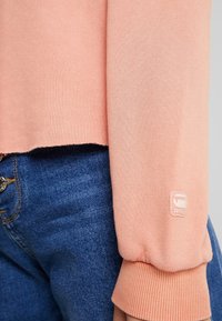 Suéter rosa claro con puños acanalados, diseño recortado y textura suave. Presenta un parche con el logo en la manga. Combinado con jeans de mezclilla azules.