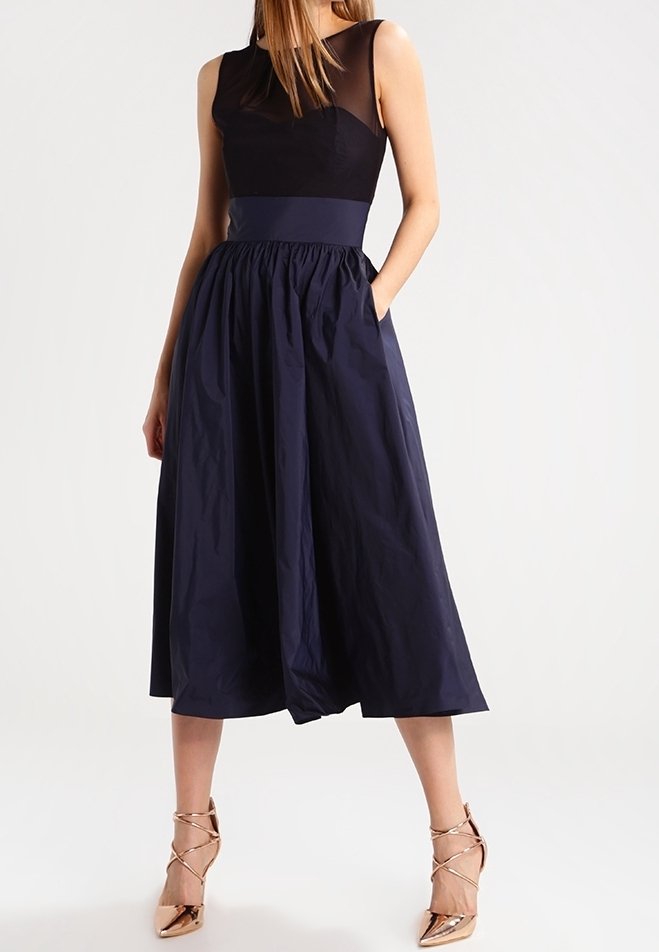 Femme portant une robe midi sans manches noire et bleu marine avec un haut transparent et des chaussures à talons hauts métalliques dorées, debout avec une main dans la poche.