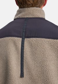 Chaqueta de forro polar beige con un panel acolchado gris oscuro en la parte superior de la espalda. Presenta una superficie texturizada y una etiqueta vertical con el logo en el centro.