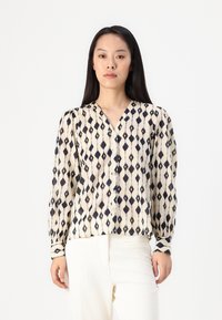 Long-sleeve blouse in cream with a navy diamond pattern, featuring a V-neckline and button front. Lightweight fabric with a smooth texture.