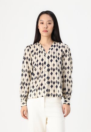 Blouse à manches longues en crème avec un motif losange bleu marine, présentant un col en V et une fermeture à boutons. Tissu léger avec une texture douce.