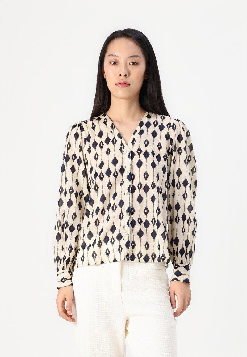 Long-sleeve blouse in cream with a navy diamond pattern, featuring a V-neckline and button front. Lightweight fabric with a smooth texture.