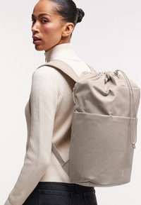 Zaino beige con coulisse, dalla texture liscia, con spallacci regolabili e una tasca con zip laterale, indossato sopra un maglione a collo alto color crema.
