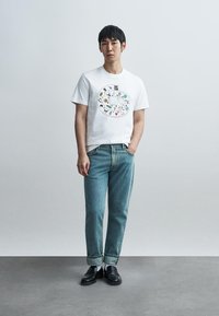 Λευκό βαμβακερό t-shirt με πολύχρωμα τυπωμένα σχέδια, συνδυασμένο με ανοιχτό μπλε τζιν παντελόνι. Μαύρα παπούτσια slip-on και λευκές κάλτσες ολοκληρώνουν την εμφάνιση.