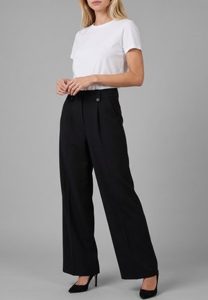 Donna che indossa una t-shirt bianca a maniche corte infilata in pantaloni neri a vita alta e gamba larga, con tacchi alti neri, in piedi su sfondo grigio.