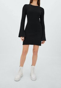 Robe noire côtelée avec de longues manches évasées, silhouette ajustée et col rond. Portée avec des bottes de combat blanches à lacets et semelles épaisses.