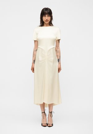 Kvinde med mørkt hår og armtatoveringer iført en cremefarvet kortærmet midikjole og sorte sandaler med høje hæle og stropper, stående oprejst.