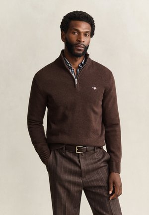 Homme barbu portant un pull marron à fermeture éclair quart sur une chemise à carreaux et un pantalon marron à fines rayures, debout avec une main dans la poche devant un fond uni.