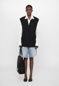 Black oversized vest with sharp lapels, paired with a white button-up shirt and frayed denim shorts. Holding a black tote bag. Black flats.