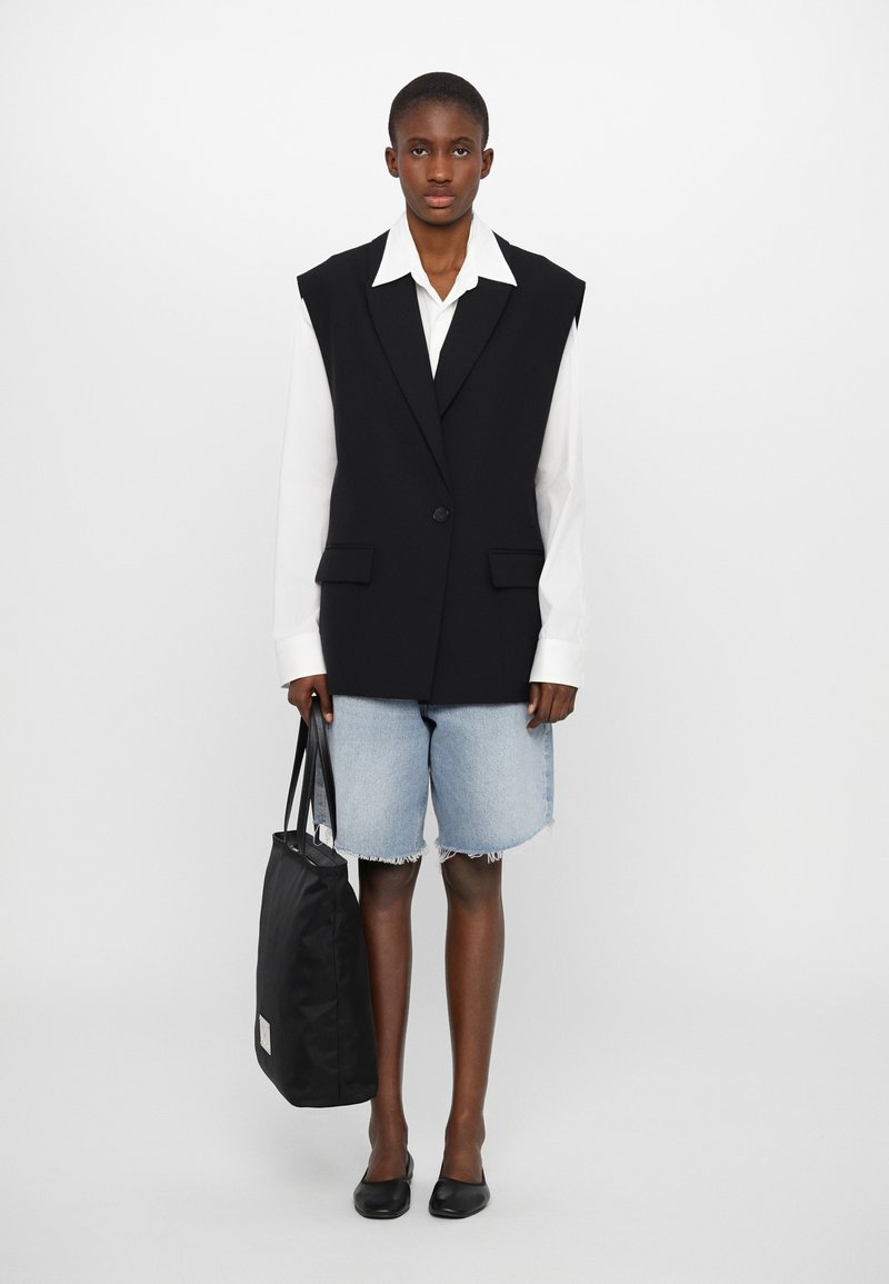 Black oversized vest with sharp lapels, paired with a white button-up shirt and frayed denim shorts. Holding a black tote bag. Black flats.
