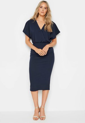 Robe midi bleu marine avec un décolleté en V, des manches courtes flottantes, une taille cintrée et une texture lisse. Portée avec des talons à brides dorées.