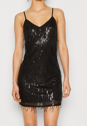 Femme portant une mini-robe noire sans manches avec fines bretelles et détails verticaux en sequins et franges, debout devant un fond uni.