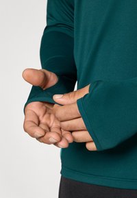 Langærmet atletiktrøje i blågrøn med tommelhuller, tætsiddende design, glat tekstur og minimal syning på ærmekanter.