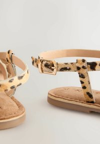 Flache Sandalen mit einem Leopardenmuster, ausgestattet mit beigen Riemen und einer goldenen Schnalle. Weicher brauner Fußbett und minimales Design.
