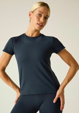 Blonde Frau trägt ein eng anliegendes schwarzes Kurzarmshirt und passende Hose, posiert mit den Händen in der Hüfte vor einem schlichten Hintergrund.