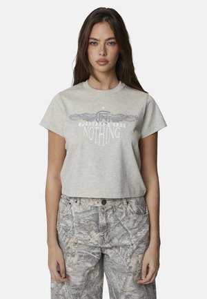 Kobieta z długimi brązowymi włosami nosi jasnoszary, krótki t-shirt ze skrzydlatym logo i napisem, zestawiony z szarymi spodniami w motyw kamuflażu.