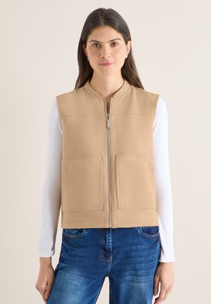Jeune femme portant une veste sans manches beige zippée sur une chemise blanche à manches longues et un jean bleu, debout devant un fond clair.