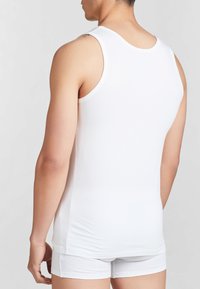 Canotta bianca di cotone con scollo rotondo, indossata sopra pantaloni corti bianchi. Il tessuto è liscio e aderente, esponendo parte di un tatuaggio sulla spalla.