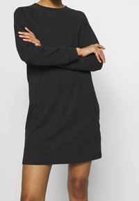 Robe sweat-shirt noire, manches longues, encolure ronde, tissu en coton lisse, coupe ample, longueur mi-cuisse, détails de couture minimalistes.
