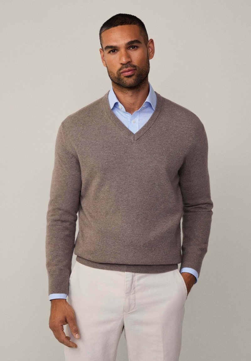 Ruskea V-pääntie villapaita, joka on valmistettu pehmeästä materiaalista, puettuna vaalean sinisen raidallisen paidan päällä. Hihoissa on kapea malli, ja villapaidassa on siisti muotoilu.