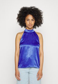 Top halter em cetim azul vibrante com um decote alto, ajuste justo e textura suave, combinado com jeans de lavagem clara.