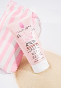 Tube de lotion bronzante instantanée étiquetée moyen-foncé, à côté d'un sac à cordon à rayures roses et blanches, sur un sable clair.