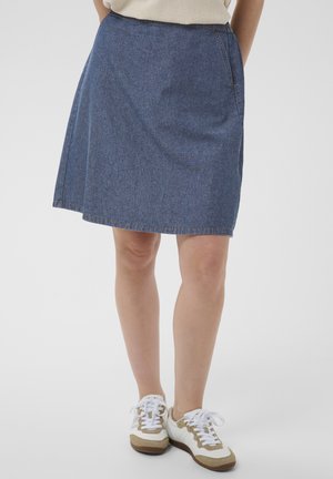 Personne portant une jupe en jean bleue longueur genou, un haut beige et des baskets blanches avec des accents marron sur un fond blanc.