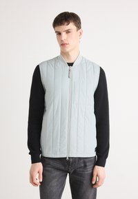 Um colete sem mangas, acolchoado e azul claro, com fecho na frente e gola canelada. Usado sobre uma camisa preta de manga longa, combinado com jeans escuros.