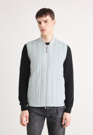 Gilet sans manches matelassé bleu clair avec une fermeture éclair devant et un col côtelé. Porté par-dessus une chemise à manches longues noire, associé à un jean foncé.