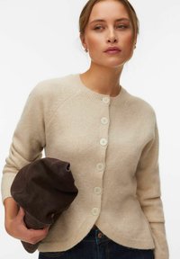 Femme portant un cardigan beige boutonné et tenant un sac en daim marron foncé plié, regardant sur le côté sur fond uni.
