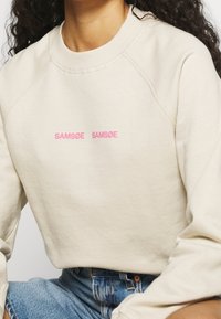 Sweat-shirt beige avec manches raglan. Présente le texte rose "SAMSNØE" imprimé deux fois sur le devant. Texture du tissu douce, coupe décontractée.
