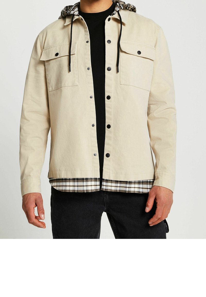 Uomo che indossa una giacca beige con bottoni neri sopra una maglietta nera e una felpa con cappuccio a quadri, abbinata a pantaloni neri, torso visibile.