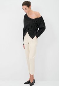 Blusa nera off-shoulder con design plissettato, dettaglio dei bottoni e maniche lunghe, abbinata a pantaloni beige su misura e scarpe nere a punta.