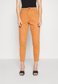 Pantalones de mezclilla beige de cintura alta con pierna ajustada, que cuentan con bolsillos frontales, cierre de botón y una textura suave. Combinados con sandalias planas negras.