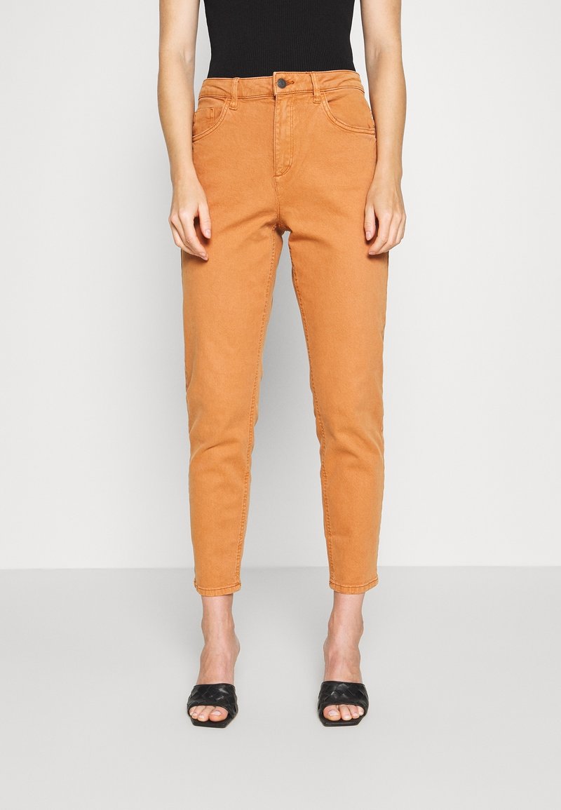 Pantalones de mezclilla beige de cintura alta con pierna ajustada, que cuentan con bolsillos frontales, cierre de botón y una textura suave. Combinados con sandalias planas negras.