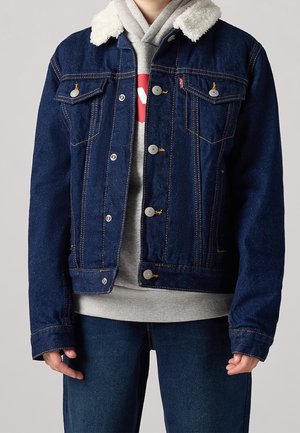 Personne portant une veste en jean bleu foncé avec un col en peau lainée blanche sur un sweat à capuche gris et un jean bleu foncé, debout devant un fond uni.