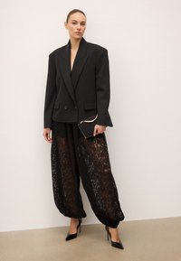 Must tumeekitsed bleiser laia õlgadega, kombineeritud läbipaistvate mustade pitspükstega ja mustade kõrge kontsaga kingadega, hoides väikest musta kotti.