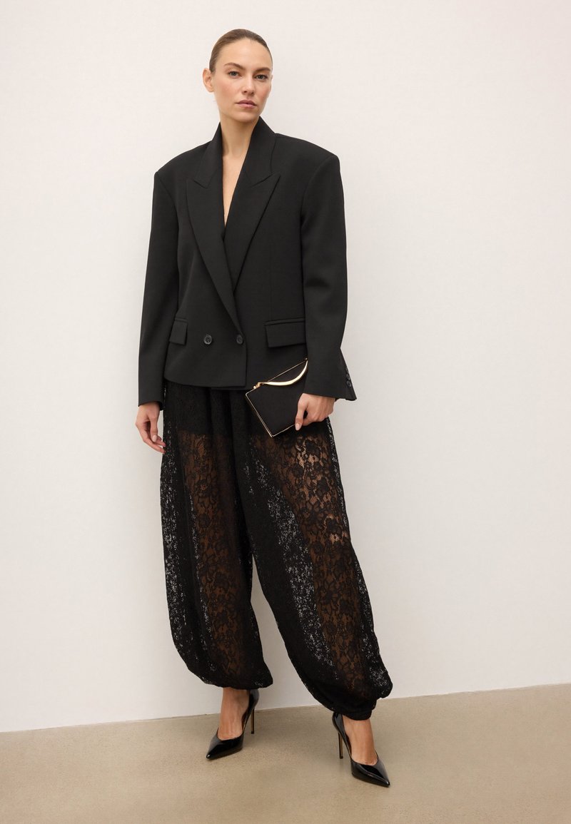 Must tumeekitsed bleiser laia õlgadega, kombineeritud läbipaistvate mustade pitspükstega ja mustade kõrge kontsaga kingadega, hoides väikest musta kotti.