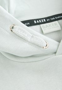 Hele-roheline kapuutsiga pusak, millel on kootud nöör ja hõbedased latitaskud. Kaelaosas musta värvi kaubamärgiga silt "BAKER by TED BAKER".