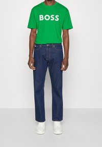 Zelené bavlněné tričko s bílým textem "BOSS", v kombinaci s modrými džíny a bílými teniskami. Ležérní střih s krátkými rukávy a rovn ými nohavicemi.