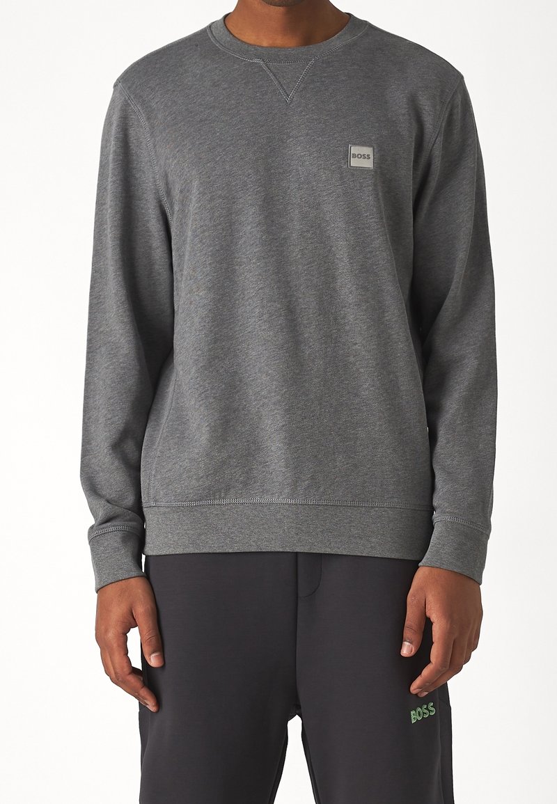 Homme portant un sweat-shirt gris uni à manches longues avec un petit logo "BOSS" sur la poitrine et un pantalon noir avec un logo vert "BOSS".