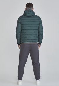Mörkgrön puffjacka med huva, med quiltad textur och elastiska manschetter, kombinerad med grå sweatpants och vita sneakers.